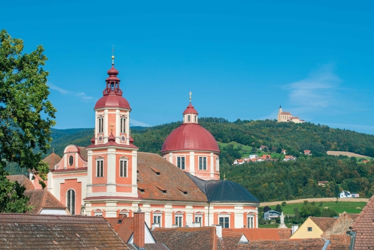 Die orange Kirche mit rotem Dach in Pöllau, im Hintergrund sieht man die gotische Kirche am Pöllauberg