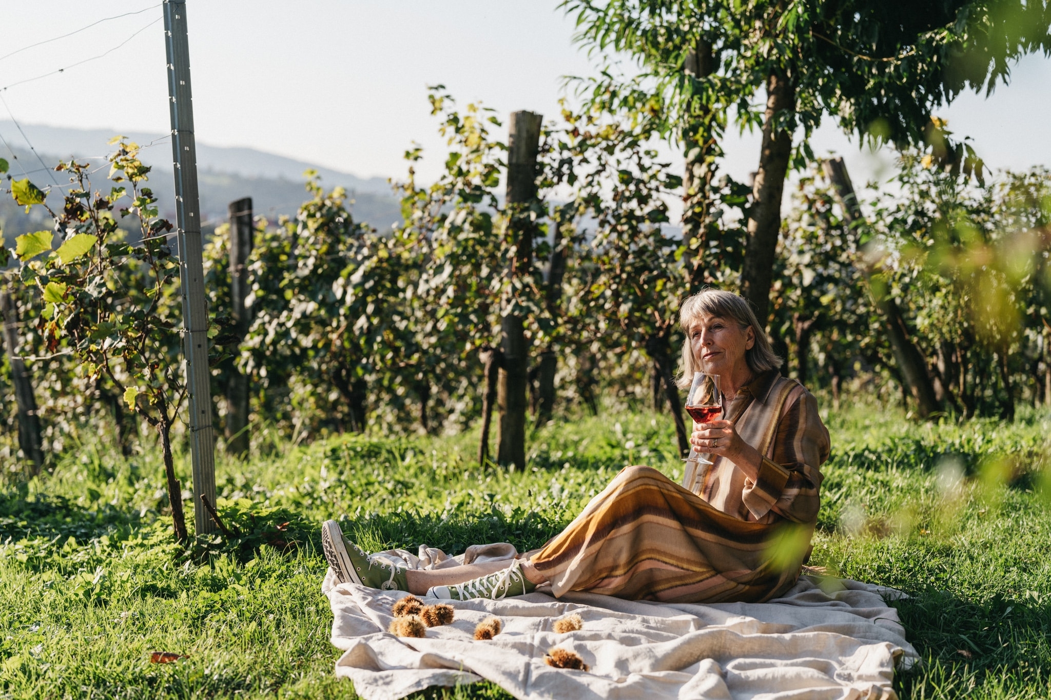 Eine ältere Frau sitzt auf einer Decke in den Weinhängen und hält ein Glas Wein in der Hand.