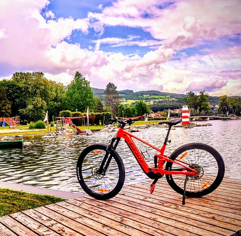Ein rotes E-Bike steht auf einem Holzsteg. Im Hintergrund ist ein See zu sehen und grüne Wiesen.